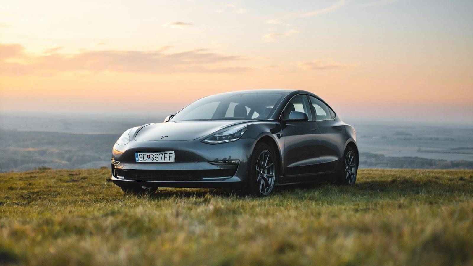 A dark grey Tesla Model 3 parked on open ground at sunrise